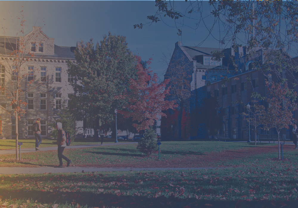A person walks across a campus lawn with autumn-colored trees, near classical university buildings under a dusky sky.
