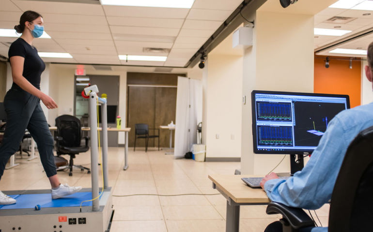 A person wearing a face mask walks on a treadmill while another person watches a computer screen displaying graphs and a 3D model.