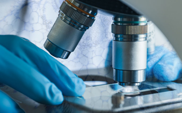 in the foreground, blue gloved hands holding a slide steady underneath a microscope. A semi-transparent overlay of microscopic cells has been transposed over the photo background
