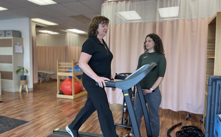 female patient is walking on a treadmill while a hospital suport staff member looks on. They are in a clinical room with exercise tools and pink hospital curtains.
