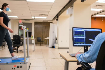 A person wearing a face mask walks on a treadmill while another person watches a computer screen displaying graphs and a 3D model.