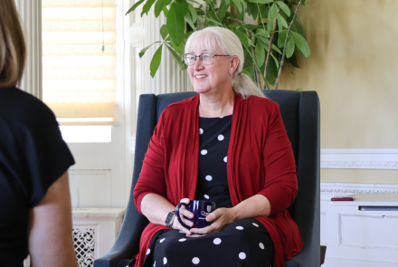 Dean Lisa Tannock sitting in a chair, holding a coffee cup in her lap and looking off to the side talking with someone