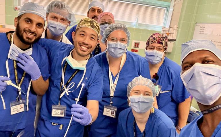 Nine doctors and nurses in scrubs, hair nets, masks and glove pose in a 'selfie' with one physician showing the pin that was removed from a patient's lung