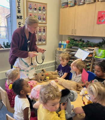 A blonde, female nursing student holds a stethoscope over a table of children sitting around her with stuffed animals.