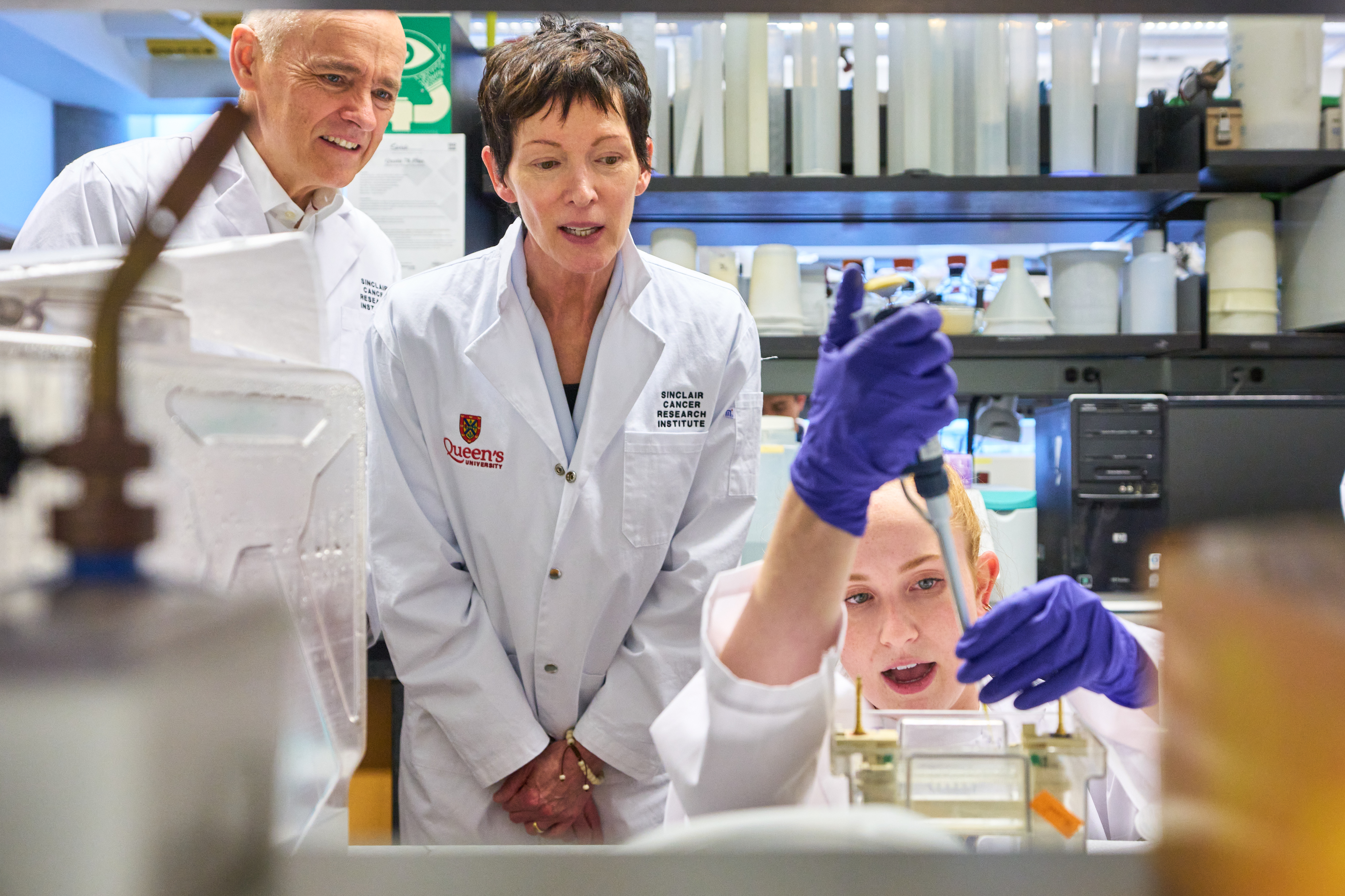 Murray and Cara Sinclair visit an SCRI laboratory, where a cancer researcher demonstrates research work to them. All three are wearing white lab coats.