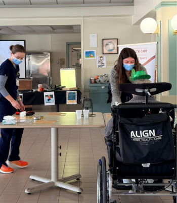 A female nursing student in scrubs and a mask is standing next to a table, while another masked female nursing student is assisting someone in a wheelchair, wearing a green hat, whose back is to the camera