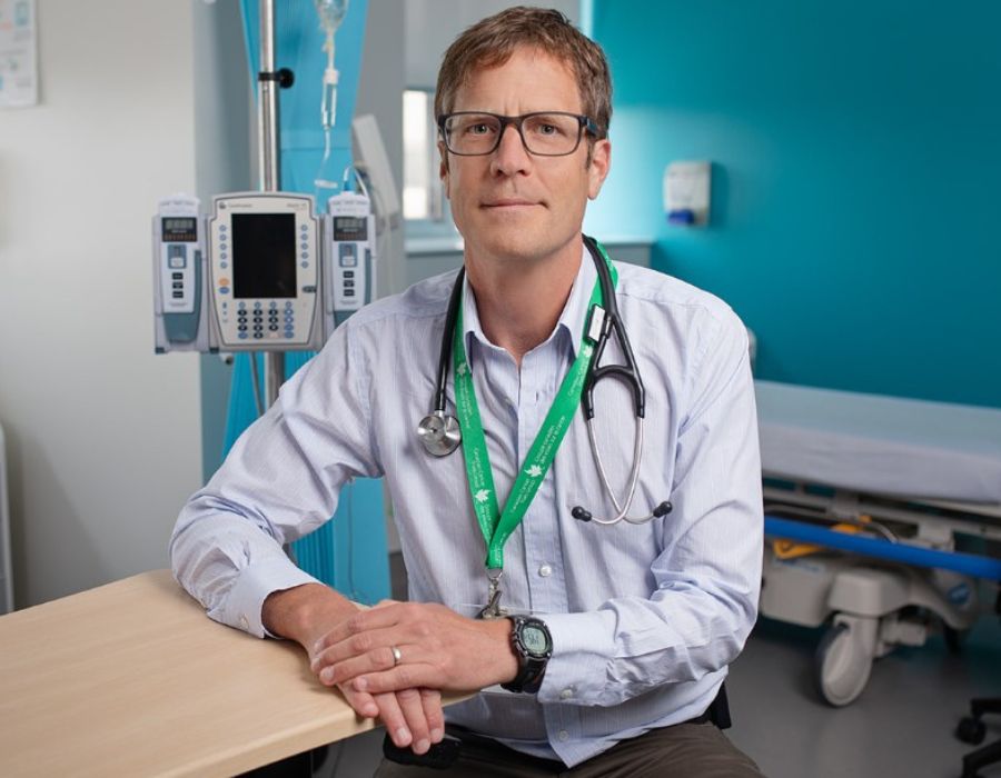 Dr. Booth wearing glasses, sitting in a chair in a clinic room, right arm resting on a table. He is staring directly into the camera