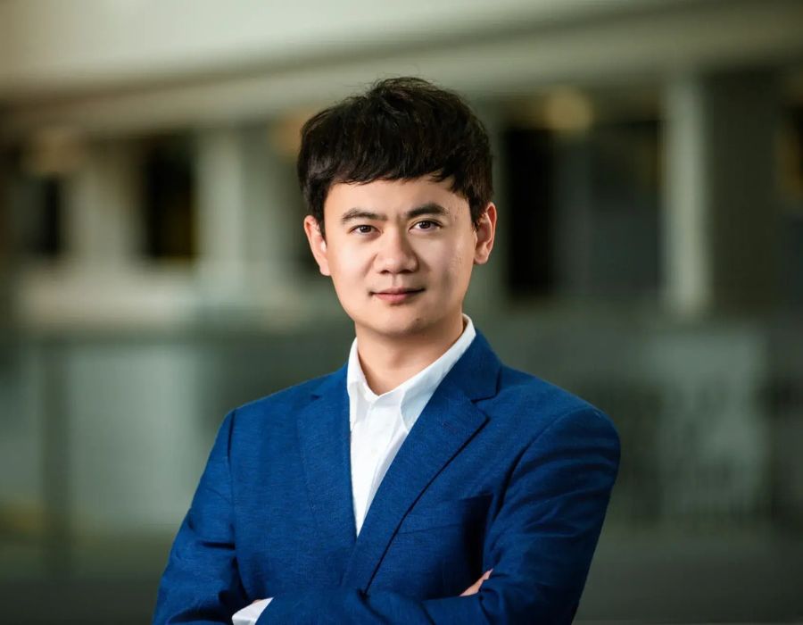 Headshot of Dr. Wang, wearing a blue suit and crisp white button-up shirt. His arms are crossed and he is staring directly into the camera.
