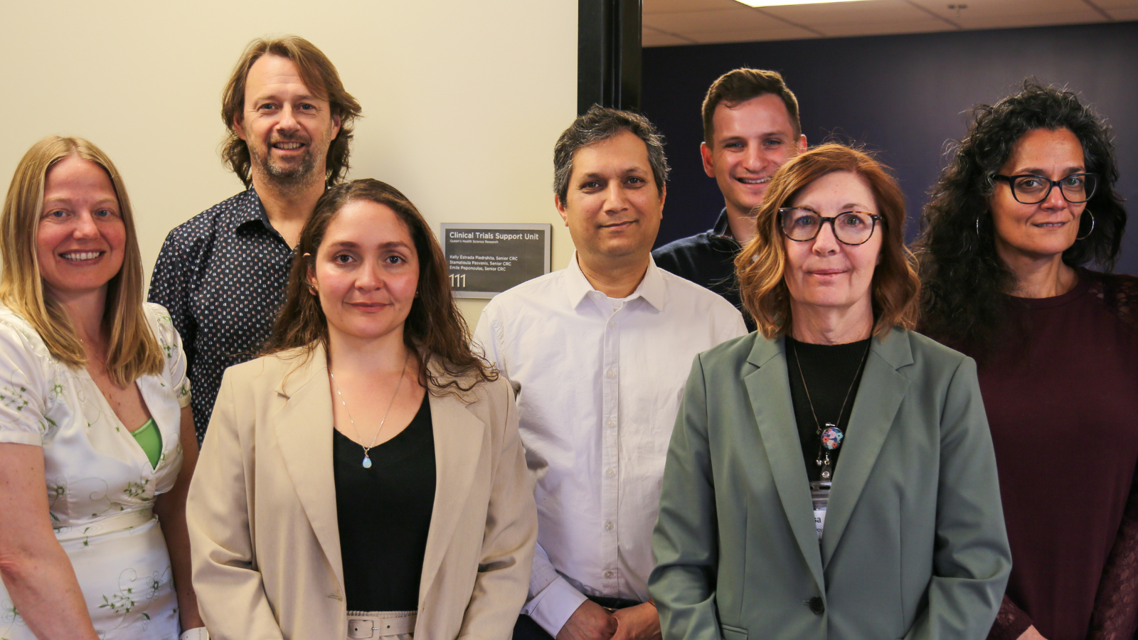 a diverse group of men and women standing together for a group photo, smiling at the camera in front of a plaque that reads 'Clinical Trials Support Unit'