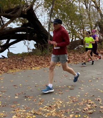 A side profile view of Dennis Christopher running past the camera during a competitive race. He is wearing a baseball cap, red hoodie sweater, grey shorts and blue running shoes.