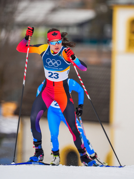 Alison Mackie racing on cross country skis, wearing an olympic games sports pinney with the number 23. She's got polarized sunglasses on and there are other ski racers behind her in the background.