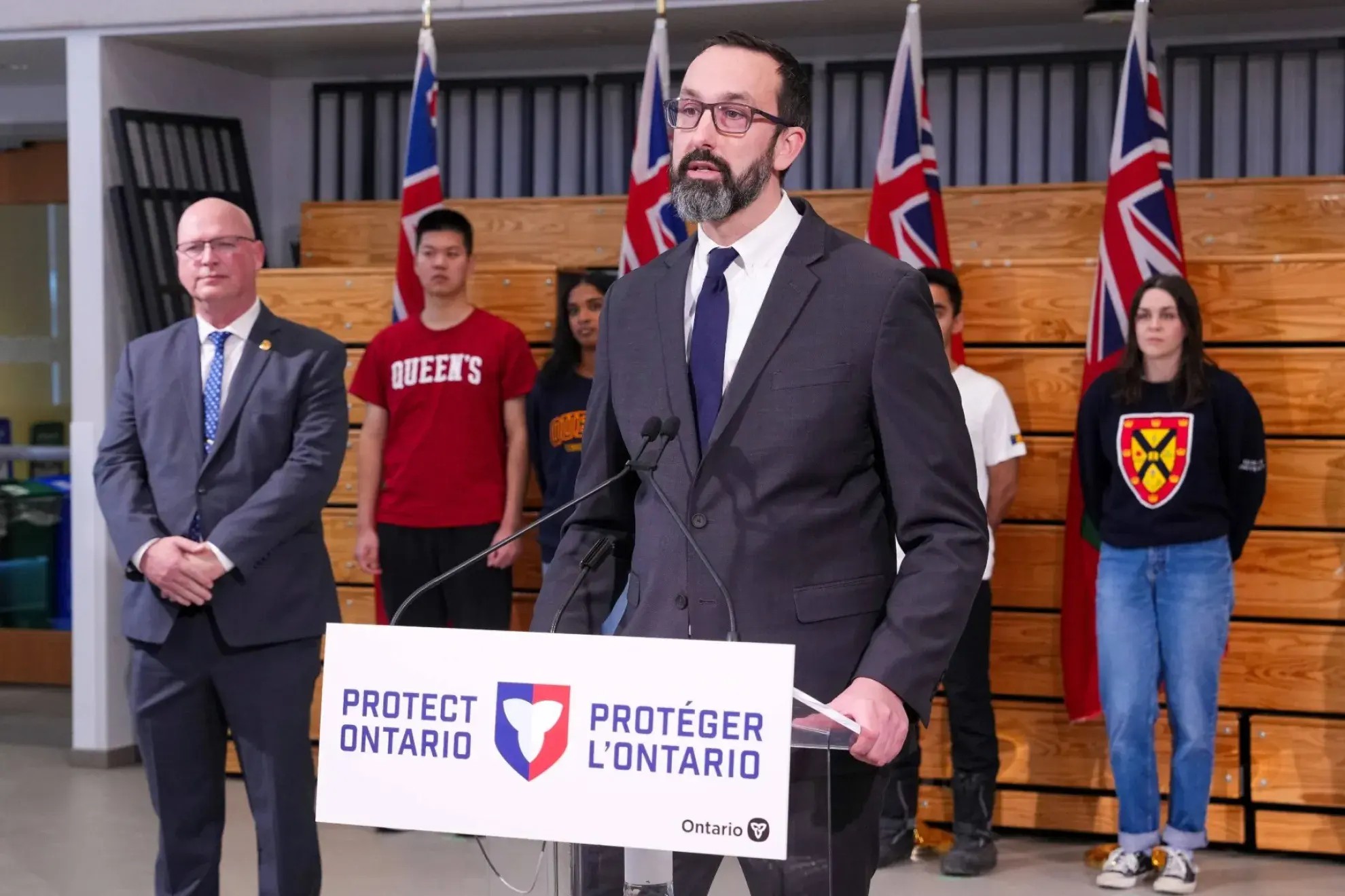 Minister Nolan Quinn in a suit and tie standing at a podium that says: "Protect Ontario". Behind him stands a diverse group of students.
