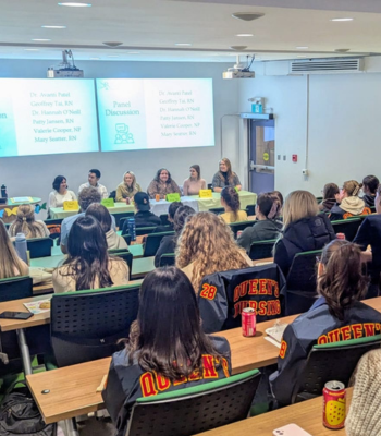 students sitting in tiered rows of lecture hall seats, backs to the camera, facing a panel of nursing experts sitting on stage in a row