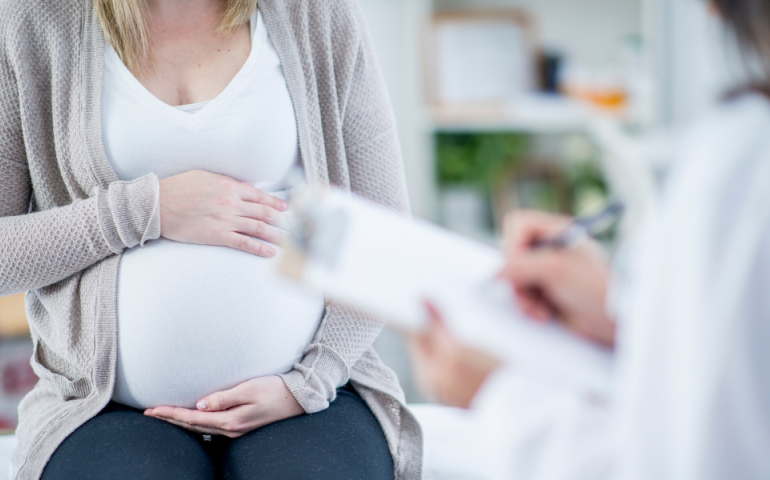 close up of a pregnant woman with her hands around her abdomen, with a health professional in the foreground filling out a form on a clipboard