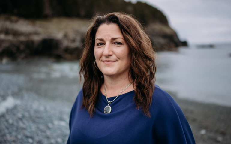 Dr. Heidi Cramm, wearing a blue sweater and necklace, long hair, smiling and standing on a rocky shoreline beach