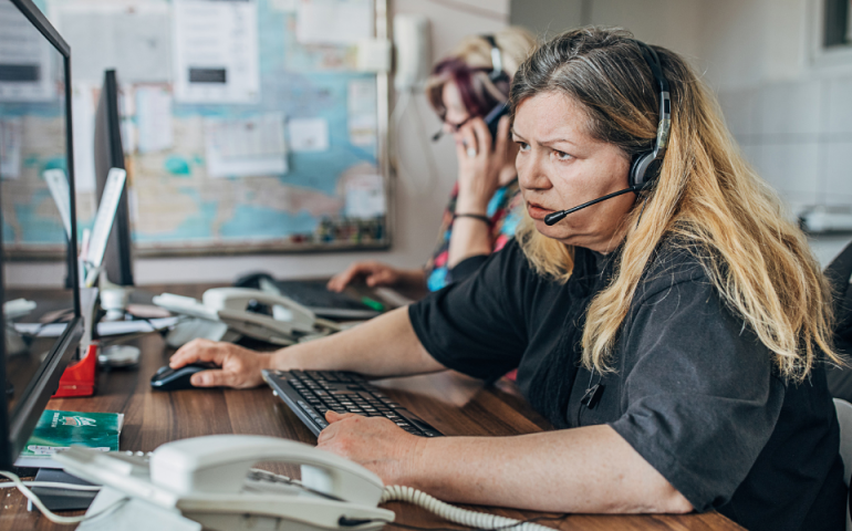911 operator sitting at a desk in front of a large computer monitor, a phone is in the foreground, she is wearing a headset and operating a computer mouse with her right hand. She looks very distressed while talking to someone on the call 