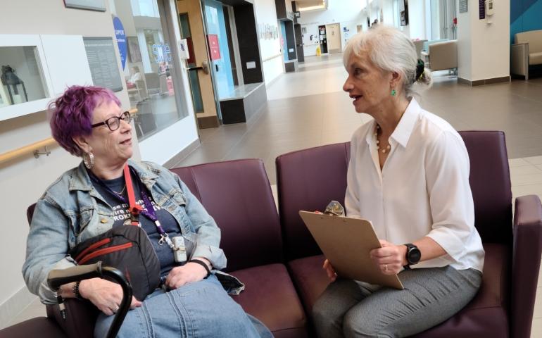 Dorothy Kessler talks to patient collaborator Isabel Guggenheimer, sitting on chairs and facing each other, Dorothy has a clipboard and is wearing a white shirt and grey pants, Isabel is wearing a denim skirt and jacket and holding a cane. Behind them is a large hallway and hospital lobby area
