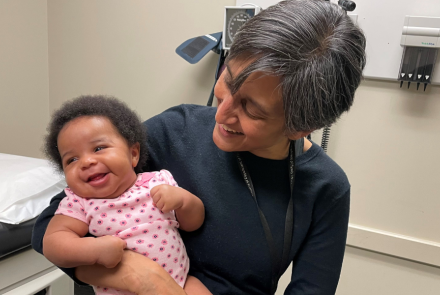 healthcare professional Dr. Rupa Patel holding a smiling baby in a medical examination room.