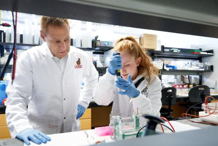 Dr. Paul Kubes wearing a lab coat and gloves looks on as a student wearing a lab coat and gloves performs work in the lab