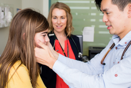 A physician conducts a routine ear and throat examination for a young patient, while a healthcare team member observes in the background.