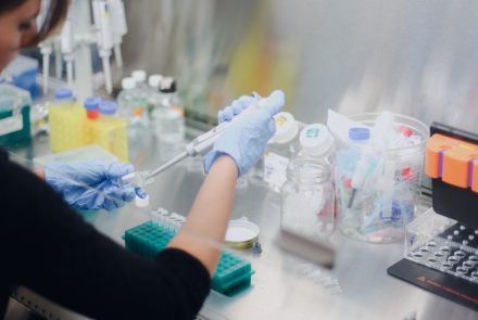 a woman with dark hair pulled back sits at a lab table, inserting a scientific device into a glass medical vial. Around her on the table are varius lab containers and equipment