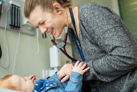 "A white female family doctor with dark blonde hair in a ponytail leans over a toddler lying on an exam table, smiling as she listens to his heartbeat with a stethoscope.