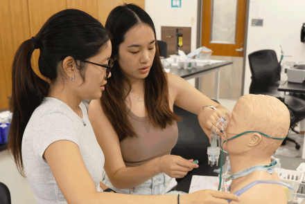 Two nursing students practice a procedure on a mannequin head and torso, with medical equipment on the table.