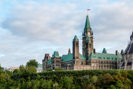 Canadian parliament buildings in full view behind lush green trees. The sky is clear and blue, there is a Canadian flag flying from the building's clock tower.