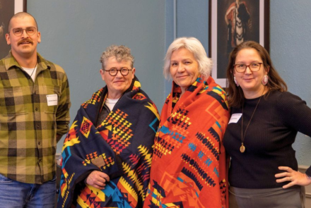 Nathan Cheechoo, Nancy Tatham, Donna Henderson, and Dr. Sarah Funnell, standing together and smiling directly into the camera. Nancy and Donna are wrapped in colorful, patterned ceremonial blankets and framed portaits of Indigenious leaders hang on the walls behind them.