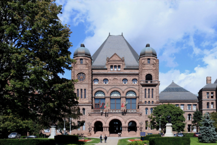 The Ontario Legislative Building with a large tree in the foreground and an Ontario flag flying from a flagpole.