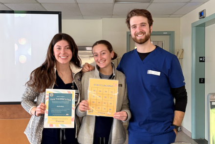 Two female and one male nursing students in medical scrubs stand behind a table holding singage for an event. The table displays a chest X-ray, a blood pressure monitor, and certificates. They are smiling at the camera.