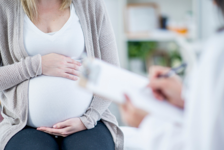 close up of a pregnant woman with her hands around her abdomen, with a health professional in the foreground filling out a form on a clipboard