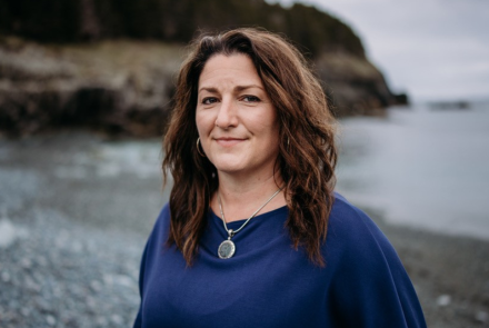 Dr. Heidi Cramm, wearing a blue sweater and necklace, long hair, smiling and standing on a rocky shoreline beach