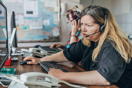 911 operator sitting at a desk in front of a large computer monitor, a phone is in the foreground, she is wearing a headset and operating a computer mouse with her right hand. She looks very distressed while talking to someone on the call 
