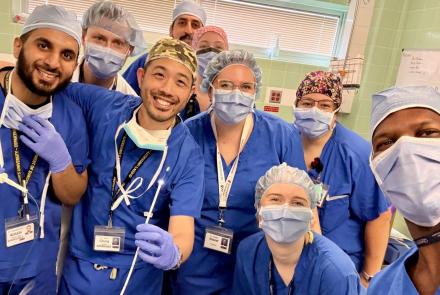 Nine doctors and nurses in scrubs, hair nets, masks and glove pose in a 'selfie' with one physician showing the pin that was removed from a patient's lung